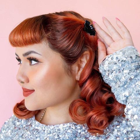 A black and silver glitter acrylic hair clip in the shape of a bat with big white eyes. Shown worn in model's updo