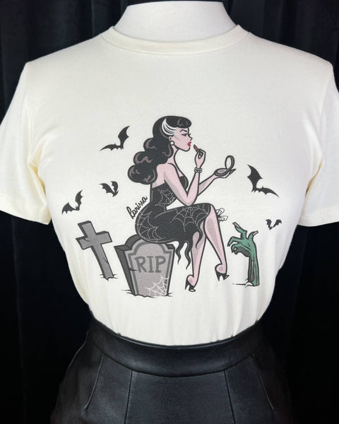 off-white fitted t-shirt with illustration of a woman sitting on a gravestone in a cemetery wearing a spiderweb patterned wiggle dress holding a compact mirror and applying lipstick surrounded by bats and a zombie hand. Shown on dress form in close up
