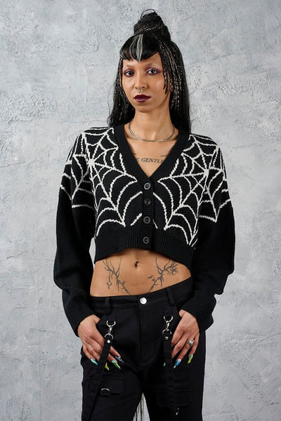 model wearing a black cropped cardigan sweater with a bright white jacquard knit-in spiderweb pattern across the chest and shoulders, featuring a V-neck and four front buttons. Shown from front