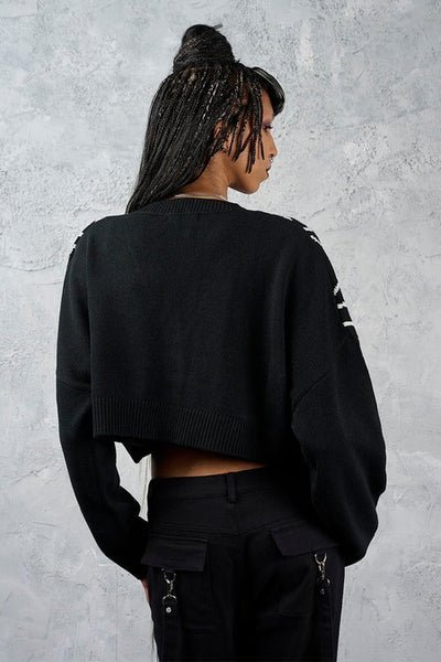 model wearing a black cropped cardigan sweater with a bright white jacquard knit-in spiderweb pattern across the chest and shoulders, featuring a V-neck and four front buttons. Shown from back