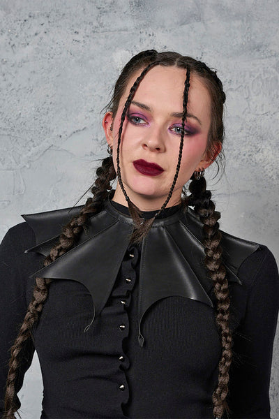 model wearing black textured faux leather collar with gunmetal tone metal snap closure in exaggerated batwing shape with scalloped edges. Shown from front