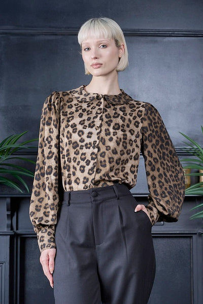 A woman with short blonde hair wearing a natural black and brown leopard print woven blouse with Peter Pan collar, slightly puffed shoulders, long balloon style sleeves, and button closure cuffs. Shown with dark charcoal gray trousers, standing in front of a dark gray paneled wall with green plants on either side.