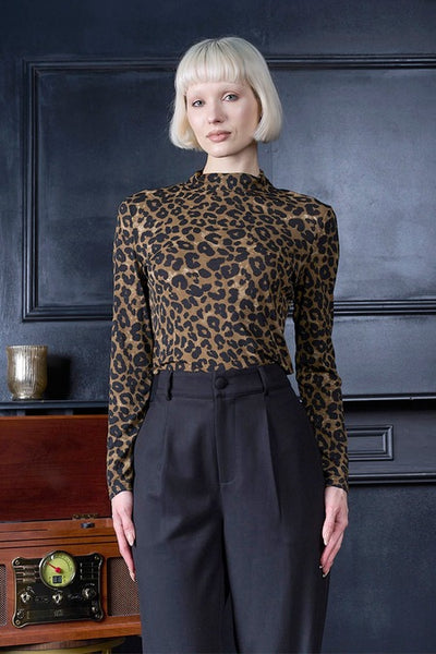 A model with a short blonde bob stands against a dark paneled wall wearing a long-sleeve leopard-print mock neck top and black high-waisted trousers.