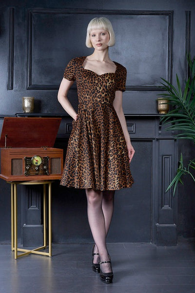 A woman with a short blonde bob stands in a vintage room wearing a brown and black leopard-print dress with short sleeves and a flared skirt. She is posed next to a wooden radio on a gold stand against a dark gray paneled wall.
