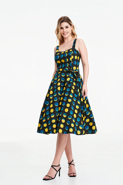 A model wearing a sleeveless black background yellow and blue moon phase print dress with a fitted sweetheart neckline princess seamed bodice with adjustable straps, removable self sash belt, swingy gathered full circle just below the knee length skirt. Shown from front