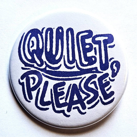 1.25” round pinback button with white background and 60s/70s style bubble lettering in white and dark blue with message ”QUIET, PLEASE”