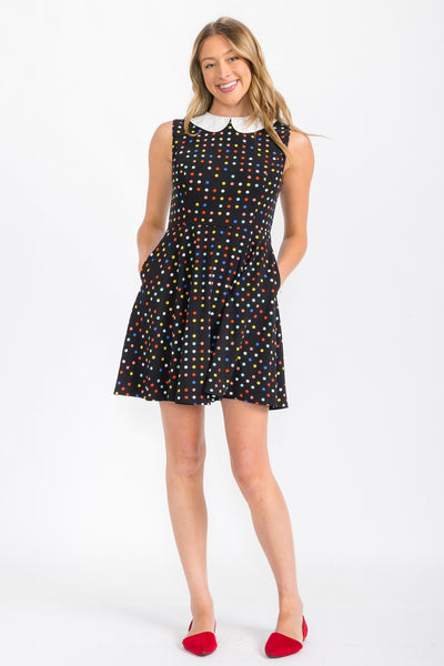 model wearing black sleeveless shift dress with pink, yellow, red, blue, and white multicolor polka dot pattern and white Peter Pan collar. Shown from front with hands in pockets