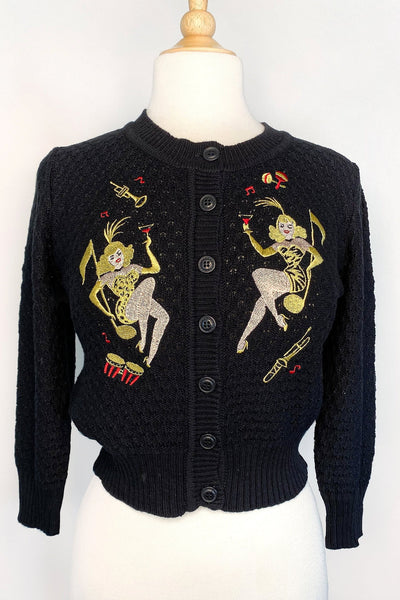 black cotton pointelle knit cardigan with embroidery detail on both sides of the chest of Vegas style yellow animal print-clad showgirls posed pinup style atop giant golden music notes surrounded by yellow and red musical instruments. Shown from the front on dress form