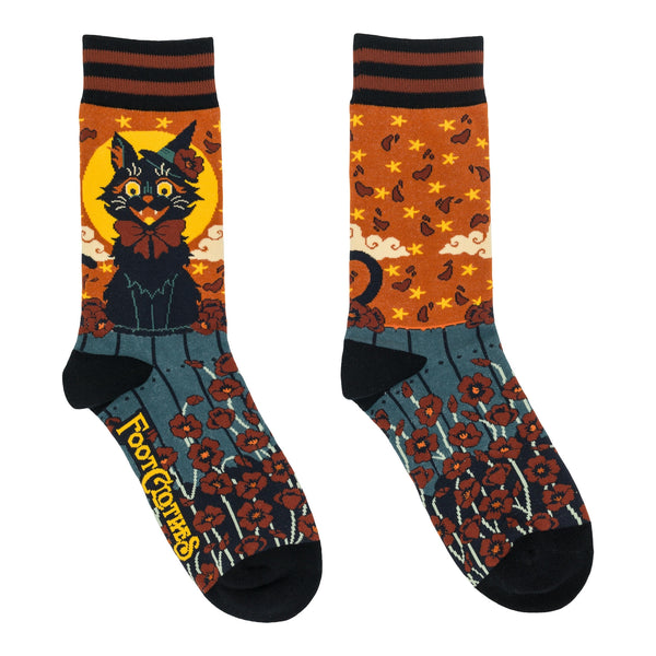 pair of unisex cotton blend crew socks with allover pattern of black witchy cat in witch hat and red bow sitting atop a wooden fence surrounded by red flowers and sky of falling red leaves and yellow stars on orange background. Shown flat