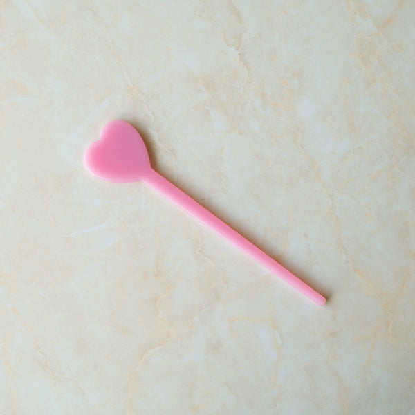 Acetate hair stick with a heart shaped topper in a shade of pink. Shown flat.