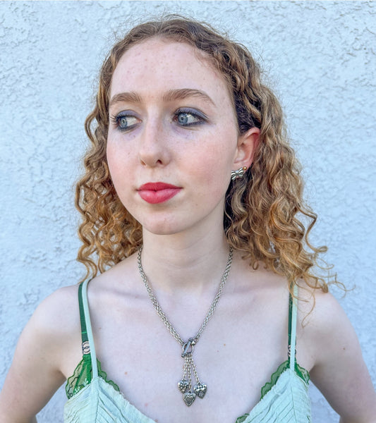 silver metal link chain toggle necklace with trio of heart charms hanging from three smaller chains at front of necklace. Worn by a model with chain doubled for shorter necklace