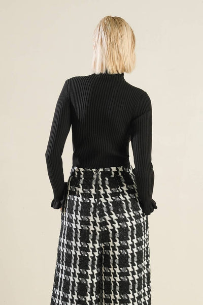 model wearing ribbed knit black mock turtleneck with long sleeves and ruffle detail at each cuff and bottom hem. Shown from the back