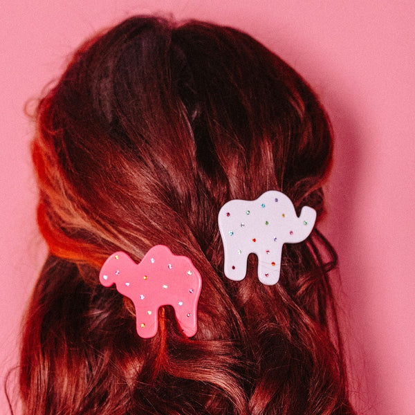 pair of acetate barrettes in the shape of a pink camel and a white elephant animal cracker, both decorated with multicolor jewels as sprinkles. Shown in updo of model with red hair from back