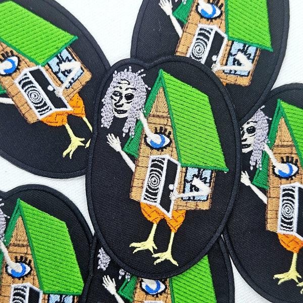 oval shaped twill embroidered patch with image of Baba Yaga and her house on chicken legs with spiral door and large eye. Shown flat with multiple patches