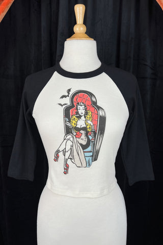 off-white crop length black raglan shoulder top with round neck, three quarter sleeves, and printed illustration of pinup style woman with stitched legs wearing gown and sitting in coffin-style large chair. Printed in red, black, blue, and yellow. Shown on dress form