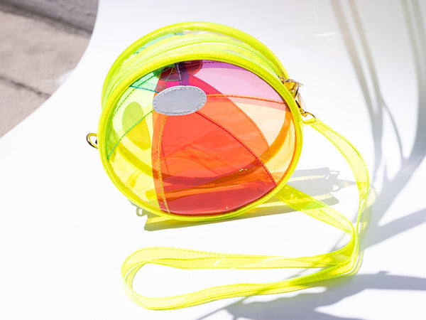bright yellow vinyl purse shaped like beach ball with red, blue, purple, green, orange, and yellow segments with zip closure top, gold metal zipper pulls and removable matching strap. Shown in sunlight from front with strap attached
