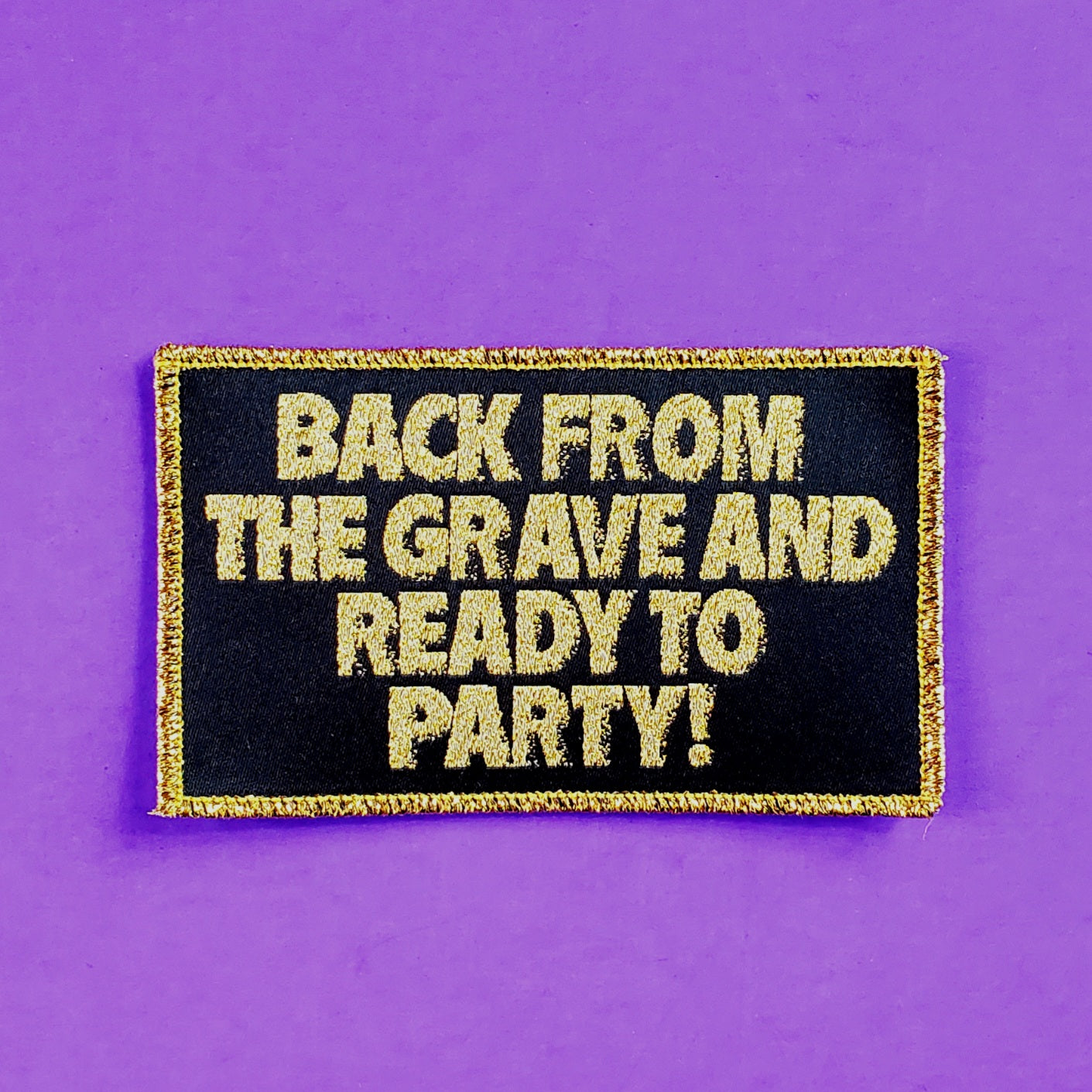 gold lamé woven patch printed with the tagline from the 80s cult zombie movie The Return of the Living Dead: “BACK FROM THE GRAVE AND READY TO PARTY!” written in gold lamé on a black background with gold lamé border