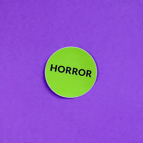 green round sticker with “HORROR” written in black capital letters, in the style of video rental store genre labels