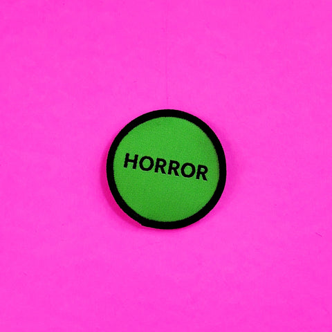 Neon green round woven patch with black border and the word “HORROR” in black all capital letters, in style of video rental store genre label sticker