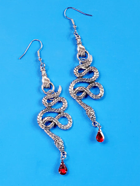 pair silver metal dangle earrings featuring a hand-shaped charm holding a stylized snake with a bright red faceted bead in the shape of a drop of blood