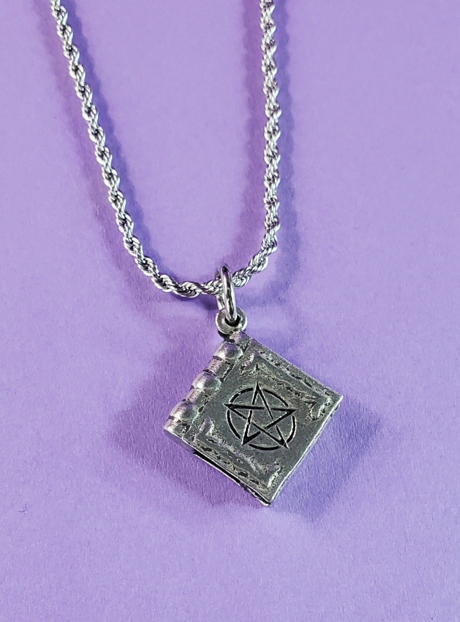 American pewter charm shaped like an ancient book of spells with a large pentagram on the front. Hanging on a stainless steel rope chain. Shown from the front