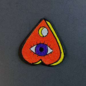 red, yellow, white, and purple embroidered patch of a planchette decorated with an all-seeing eye
