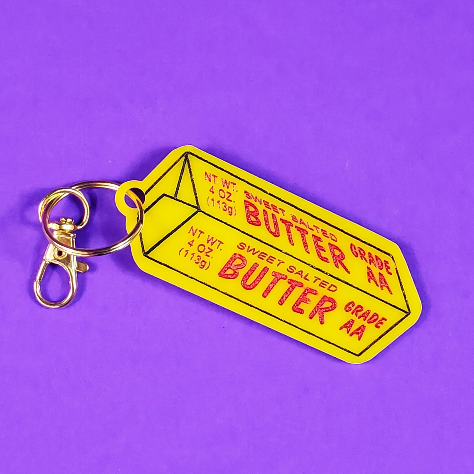 Yellow laser-cut acrylic keychain in the shape of a stick of butter with red and black painted details. Split ring and hook are shiny gold metal.