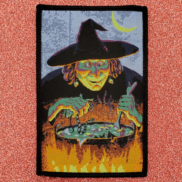 Rectangular woven patch with black embroidered border and image of a witch standing below a crescent moon above a large cauldron 