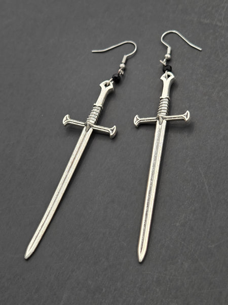Long silver metal sword charm dangle earrings with a black faceted bead connecting the sword to the fishhook ear hardware. Shown from the front