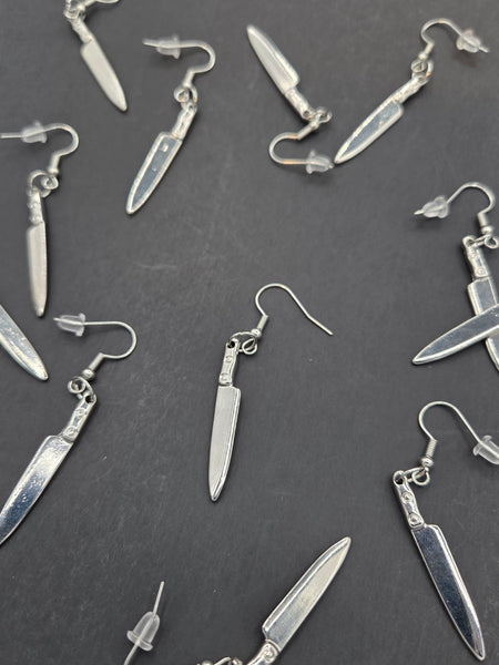 many silver metal kitchen knife shaped dangle earrings shown flatlay against a black background