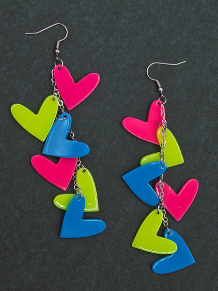 Colorful neon pink, blue, and yellow heart-shaped charms on long silver chain dangle earrings on black background