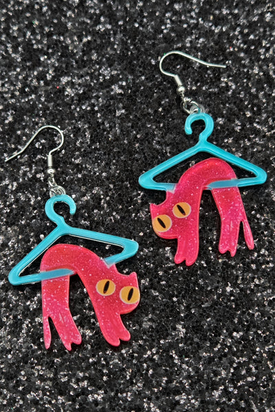 Dangle earrings made of laser-cut acrylic with printed image of pink cat designs on blue hangers against a glitter black background