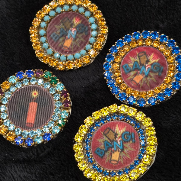 Four round lenticular dynamite/'BANG!' brooches with colorful rhinestone borders, shown on a black background