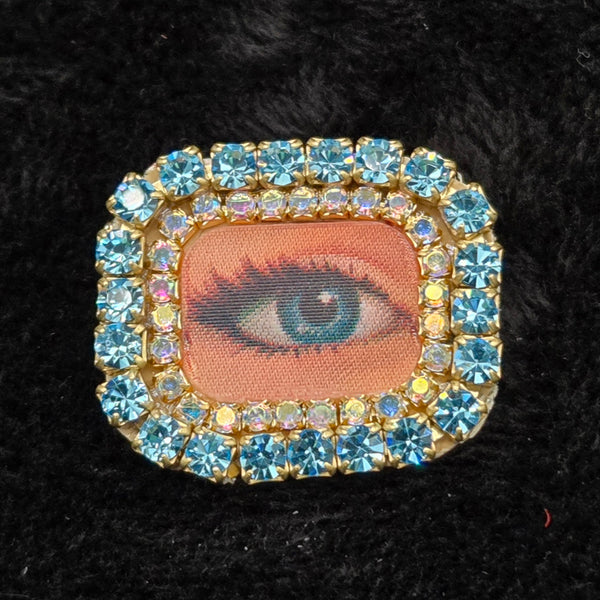 rectangular brooch with lenticular winking eye design and colorful rhinestones border, shown on a black background
