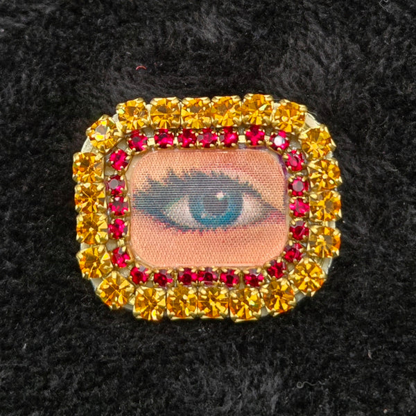 rectangular brooch with lenticular winking eye design and colorful rhinestones border, shown on a black background