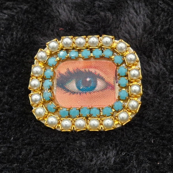 rectangular brooch with lenticular winking eye design and colorful pearl and rhinestones border, shown on a black background