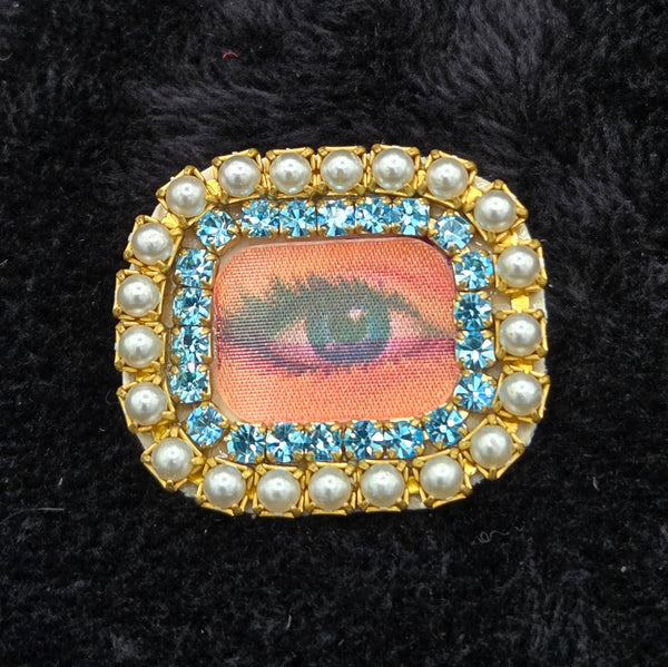 rectangular brooch with lenticular winking eye design and colorful pearl and rhinestones border, shown on a black background