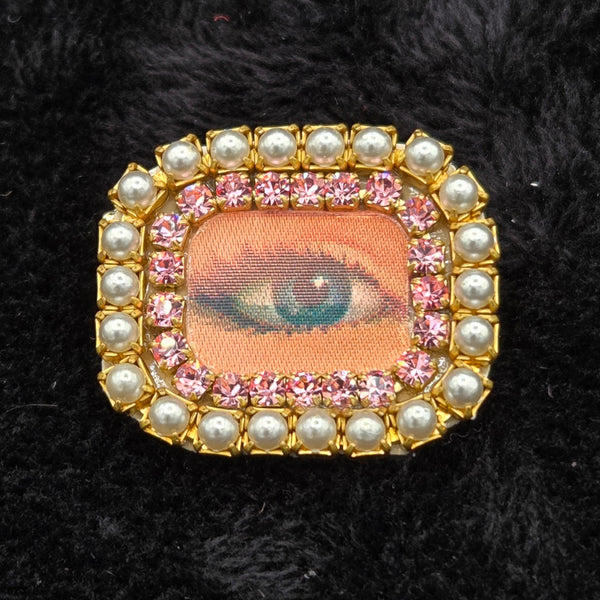 rectangular brooch with lenticular winking eye design and colorful pearl and rhinestones border, shown on a black background
