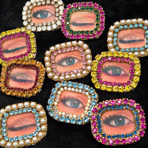 rectangular brooches with lenticular winking eye design and colorful rhinestone borders, shown on a black background