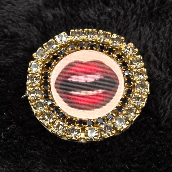 round brooch with lenticular red-lipped mouth design and rhinestone embellishments on a black background