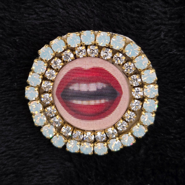 round brooch with lenticular red-lipped mouth design and rhinestone embellishments on a black background