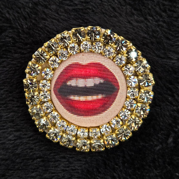 round brooch with lenticular red-lipped mouth design and rhinestone embellishments on a black background