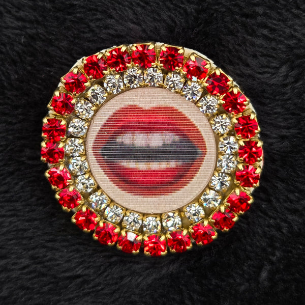 round brooch with lenticular red-lipped mouth design and rhinestone embellishments on a black background