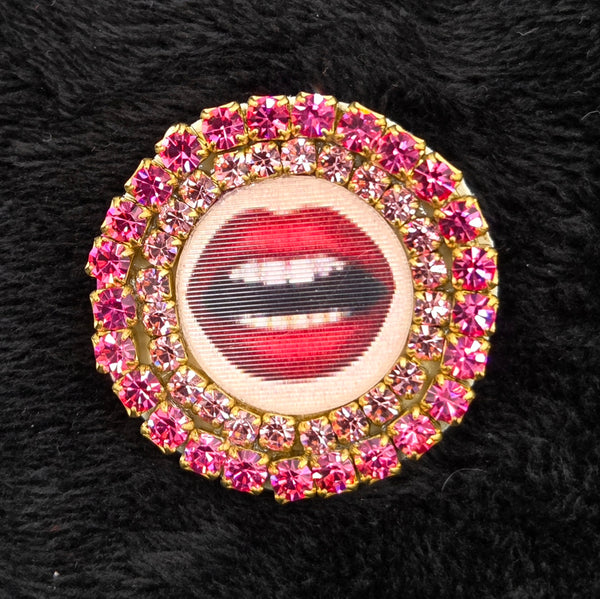 round brooch with lenticular red-lipped mouth design and rhinestone embellishments on a black background