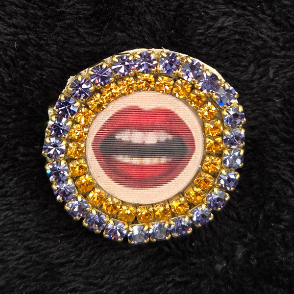 round brooch with lenticular red-lipped mouth design and rhinestone embellishments on a black background