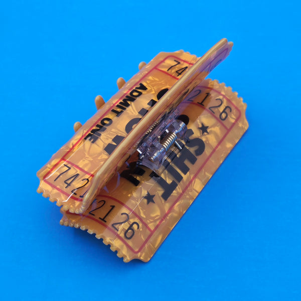 yellow marbled acetate ticket-shaped claw style hair clip with printed detail of "SHIT SHOW ADMIT ONE" shown 3/4 back view