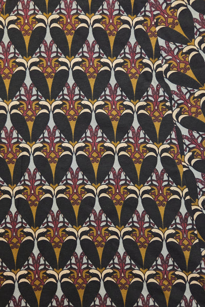 A close-up of a patterned fabric featuring a repeating Art Deco style motif with black, gold, burgundy, and cream elements on a light gray background.