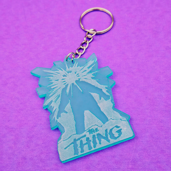 Laser-cut bright icy blue acrylic keychain with etched detail of movie poster for The Thing shown on purple background