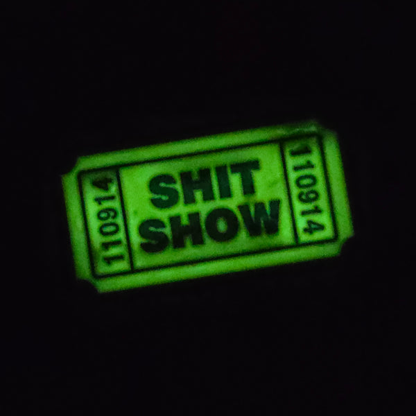 1.25" bright yellow glow-in-the-dark "Shit Show" ticket soft enameled black metal pin, shown glowing