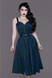 model wearing a pencil dress and swing skirt set in a deep green and blue plaid pattern. The dress has a sweetheart neckline, adjustable straps, pleated bodice with structural boning, and below the knee skirt. The skirt is a full swing skirt in matching plaid with a black faux leather strap and gold metal buckle. Shown from front